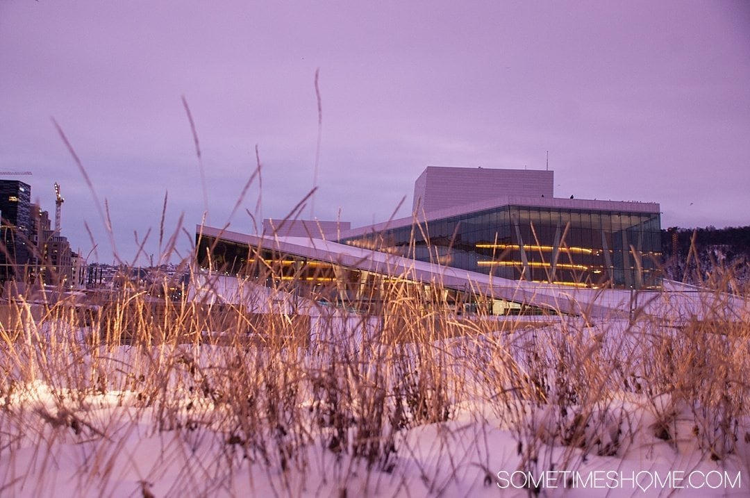 Reasons Why January is the Best Time to Visit Norway with a photo of the Opera House in Oslo during a pink sunrise, covered with snow.