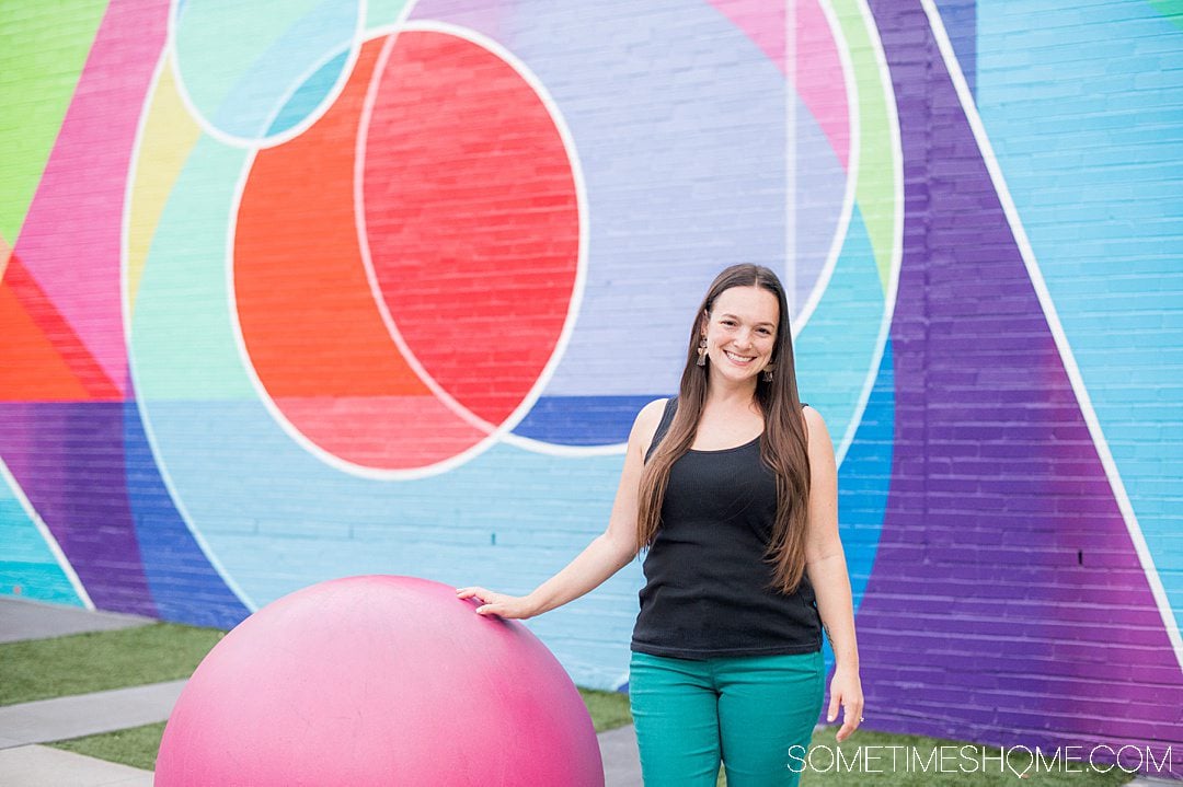 A woman standing in front of a colorful geometric mural in downtown Raleigh with various shapes including circles, triangle, and lines in red, blue, purple, green, yellow and pink