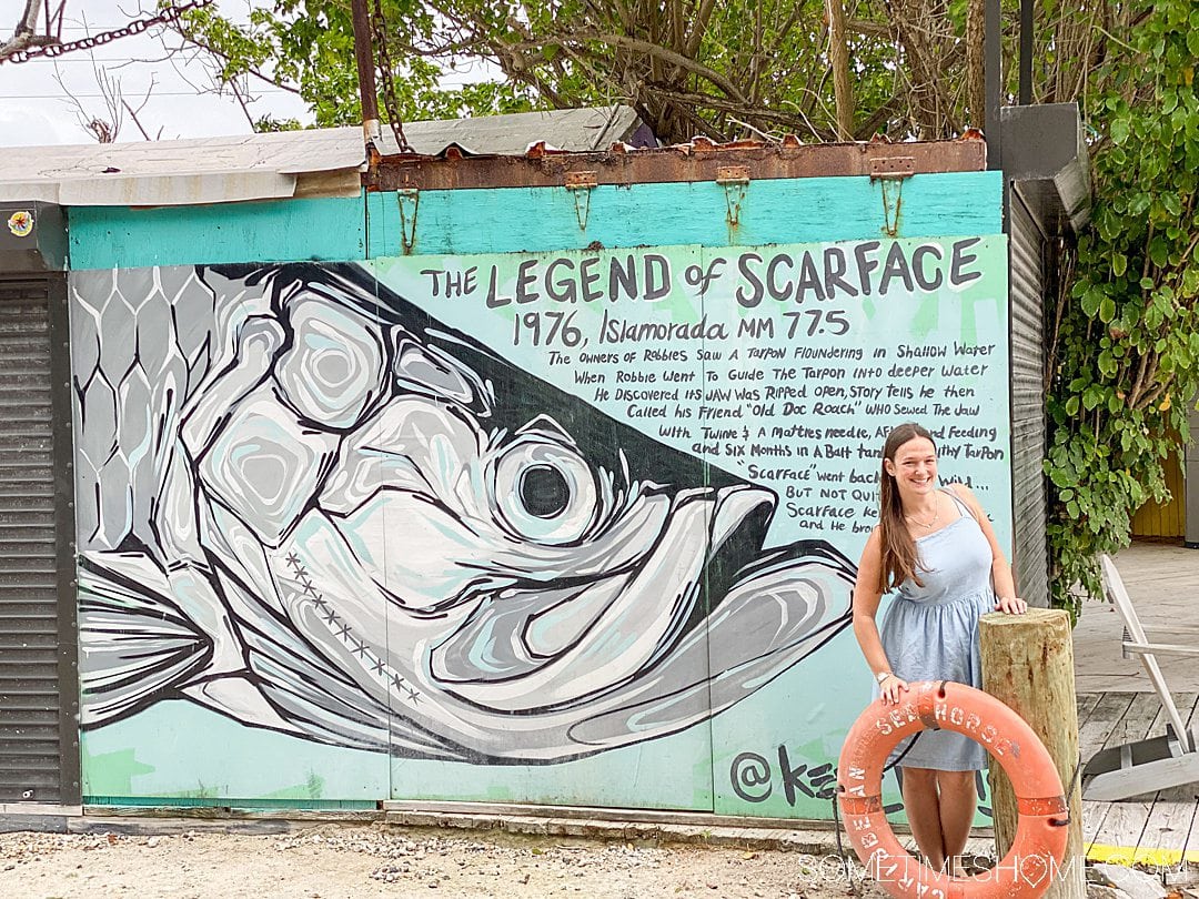 A woman posting by a life saver tube on land, with a painting of a fish behind her.