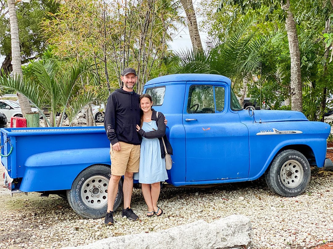 A man and a woman in front of a bright blue pick up truck with greenery behind it.