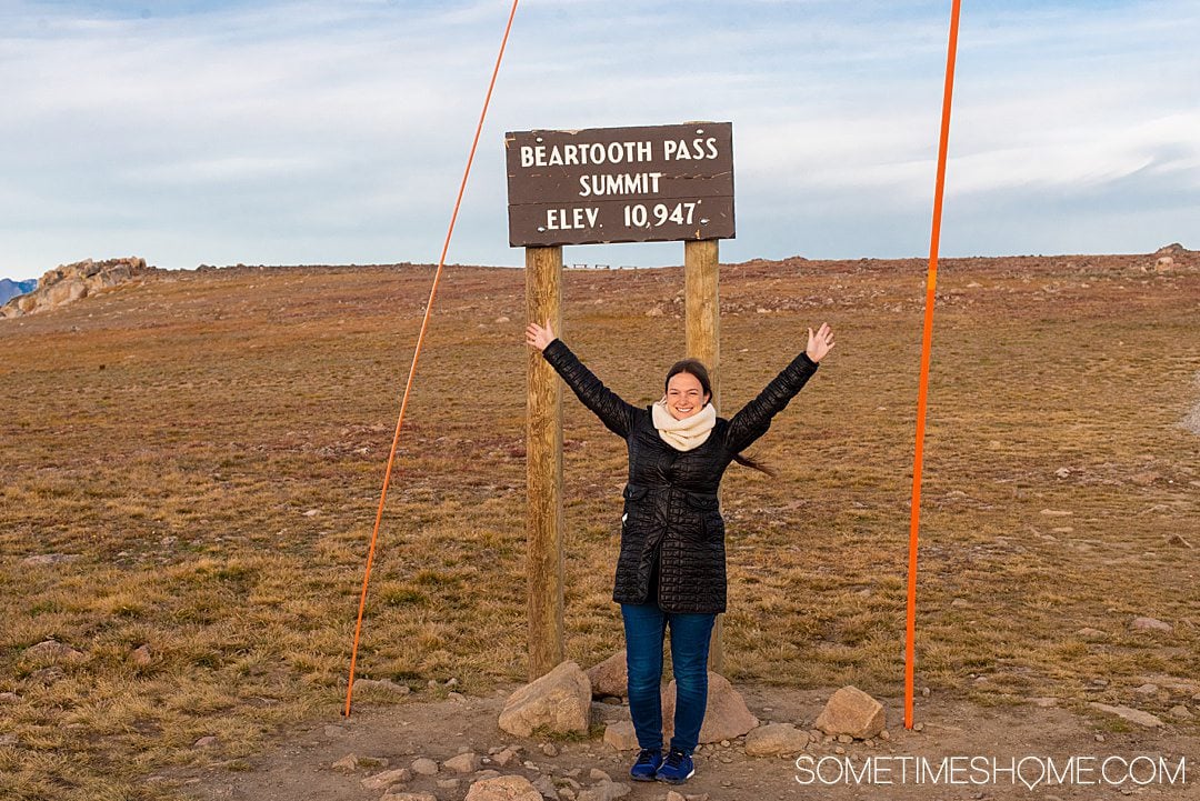 vrouw met opgeheven armen poseert voor een foto met de hoogste hoogtemarkering van de Beartooth Highway in Montana.