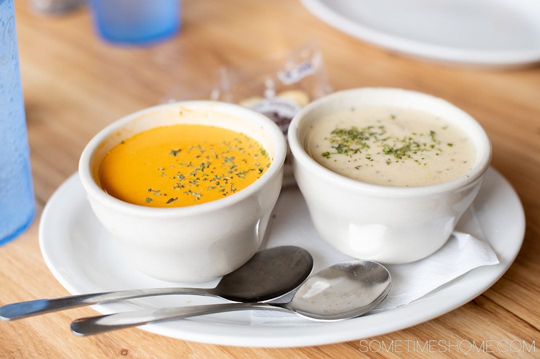 Two cups of soup, one orange and one white, on a plate with two spoons.