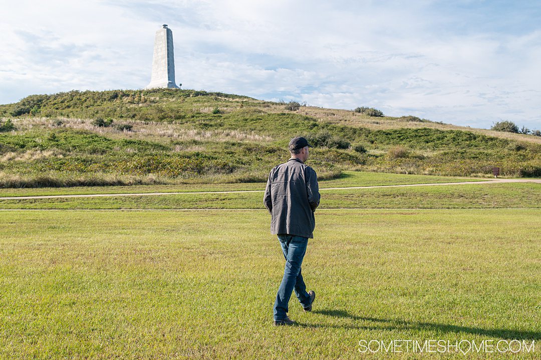 A man walking on a green lawn with a white monument on a hill in the background in the Outer Banks in October.