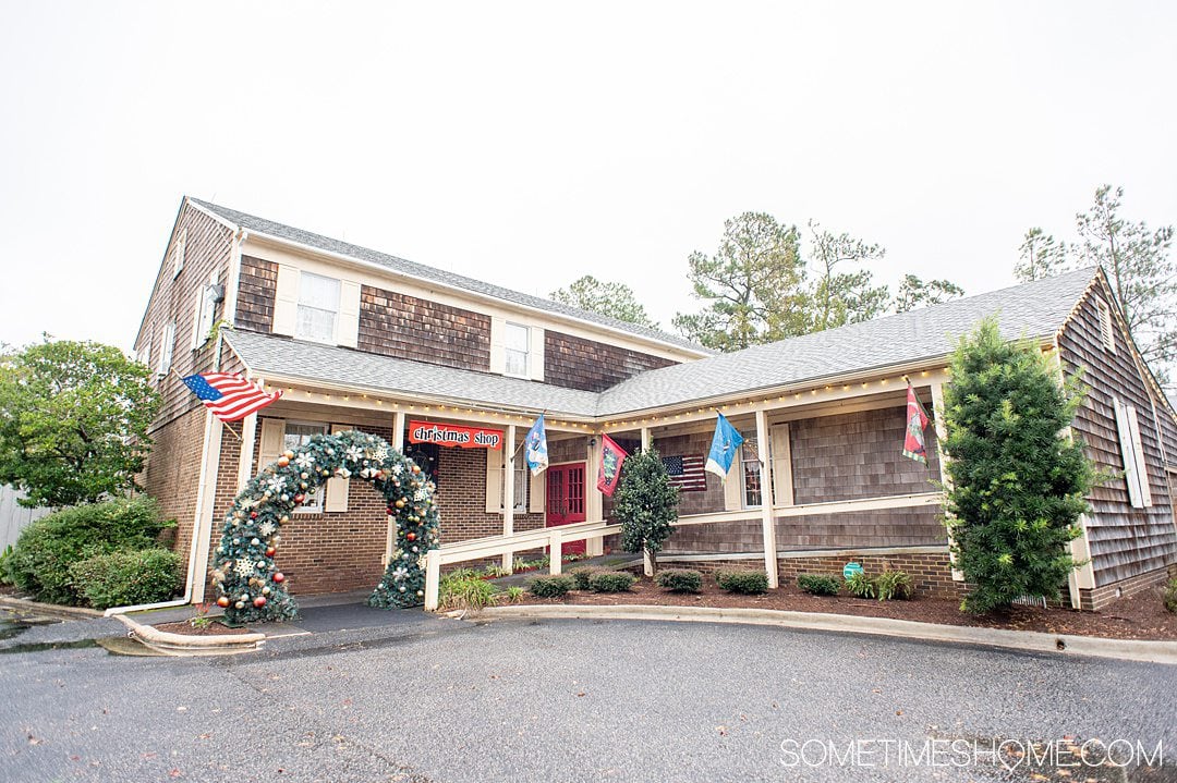 Building with an overside Christmas wreath as an entry way and porch, with wood shingles and light yellow shutters, called The Christmas Shop in the Outer Banks in NC.