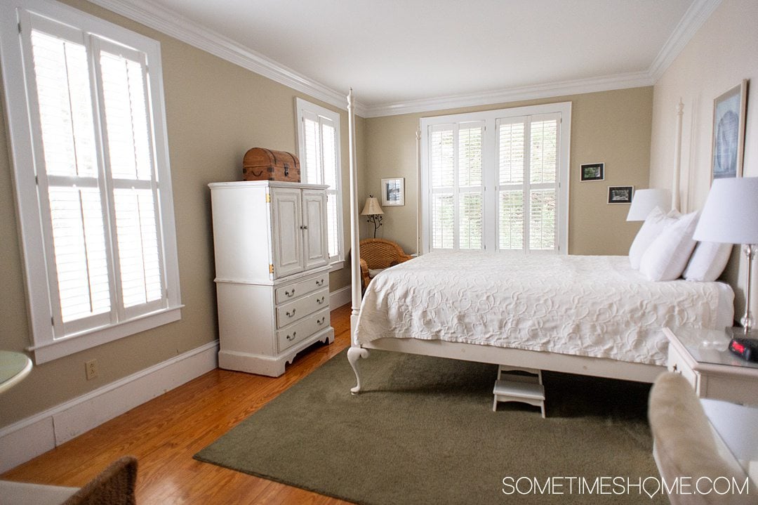 Hotel room with a white armoire on the left and white four-poster bed on the right on top of a sage green area rug at the Tranquil House Inn in the Outer Banks.