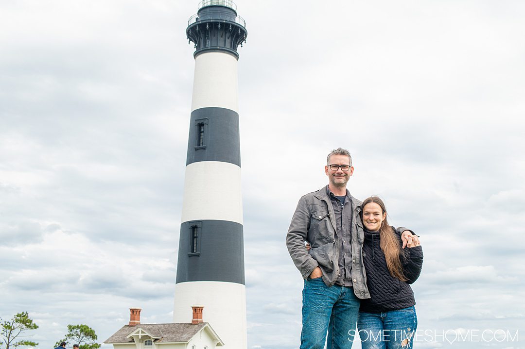 A man and a woman on the right of the photo with a black and white horizontally striped lighthouse on the left in the Outer Banks, at Cape Hatteras North Carolina.