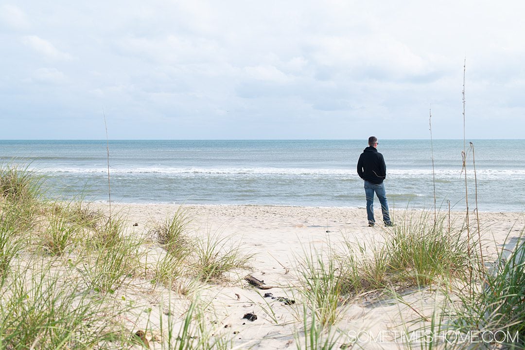 A man on the seashore at Hatteras Beach in the Outer Banks with greenery in the foreground and beach and blue sky in the background.