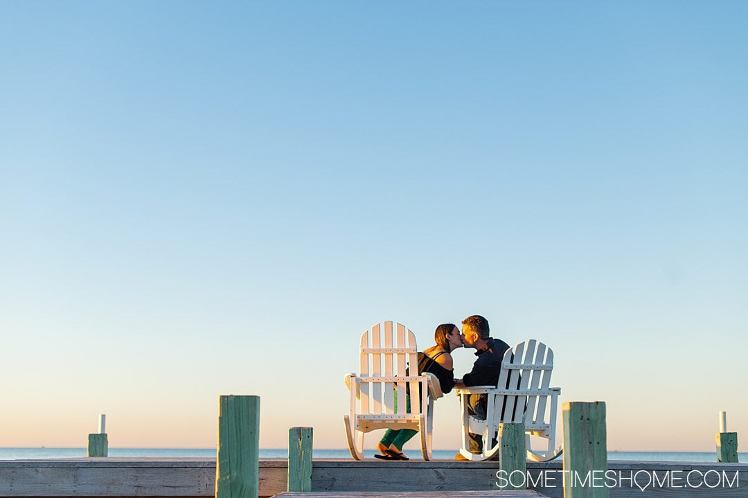 A couple kissing in white Adirondack chairs on a dock, with a blue sky fading into pink and orange during sunset at The Inn on Pamlico Sound.