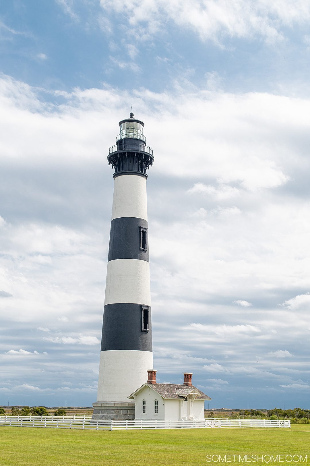 A black and white horizontally striped lighthouse in the Outer Banks in October against a blue sky with white clouds, on green grass. This lighthouse is on Cape Hatteras.