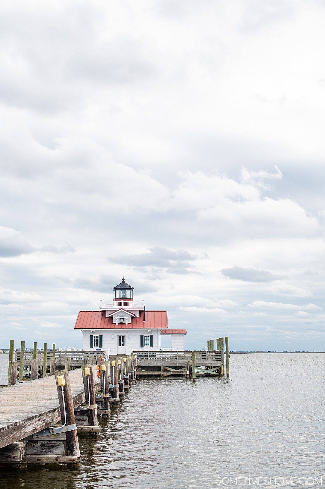 A small house at the end of the boardwalk over water and white clouds in a light blue sky. The top serves as a small lighthouse on Roanoke Island.