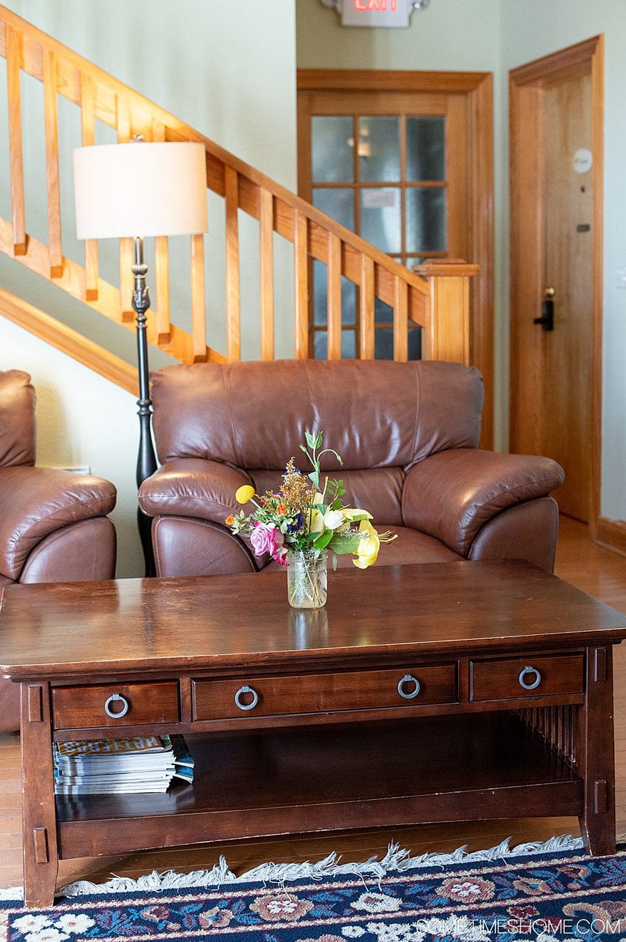 Two brown leather chairs and a brown coffee table in front of a staircase in a room. The table has a vase with colorful flowers in a room. There is a floor lamp to the left of the right chair.