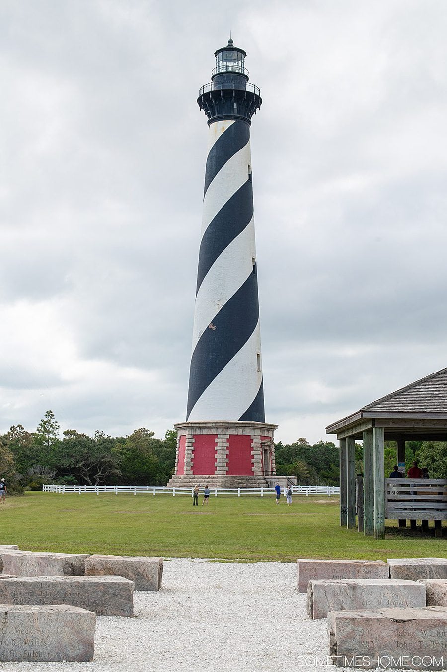 A lighthouse with a black and white swirled daymark pattern and red base, on green grass with a cloudy light grey and blue sky in Hatteras in the Outer Banks in October.