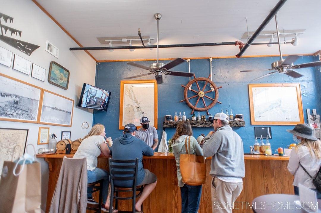 People inside at a wooden beach bar with a captain's wheel on a blue painted wall an television on the left side, at Outer Banks Distilling .