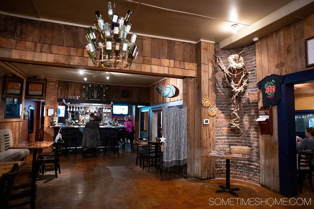 Inside of a restaurant with a wine bottle chandelier and wood finished walls and a dark floor. It's a great place to visit for one of the best Outer Banks indoor activities: drinking wine!