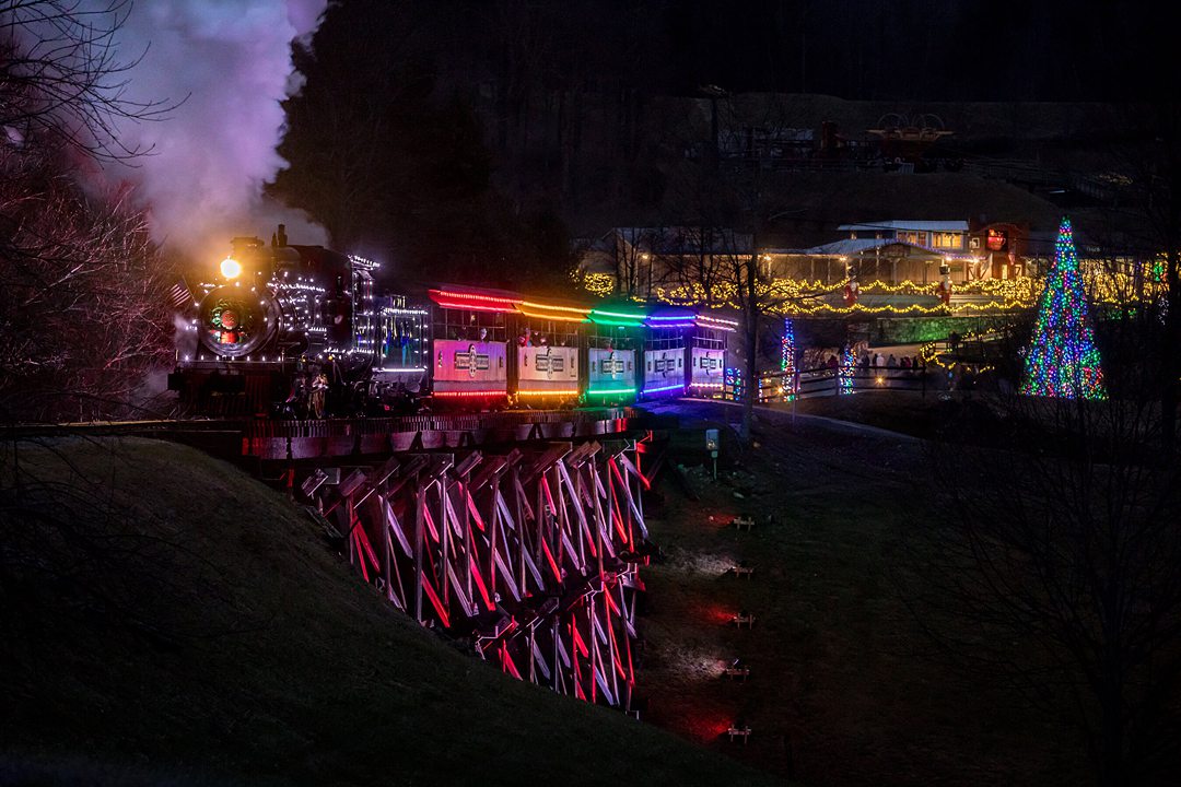 Illuminated train on a bridge above water at night with rainbow lights during a holiday winter event in NC.