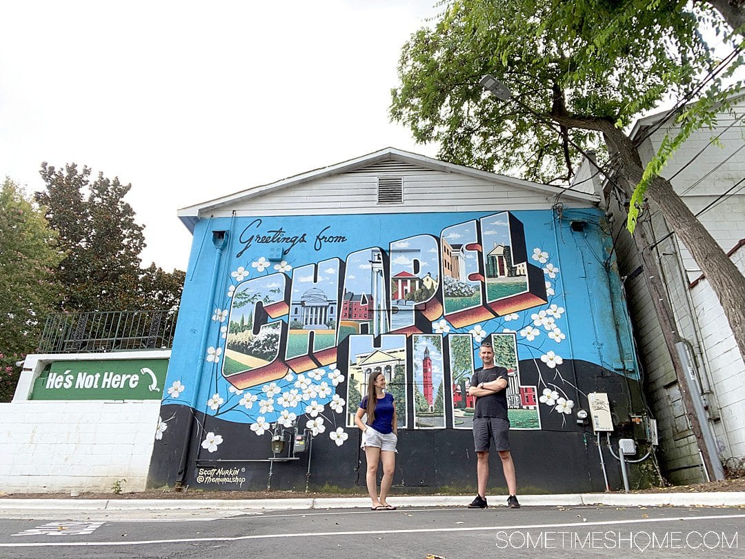 A couple in front of a colorful mural that reads, "Greetings from Chapel Hill." 