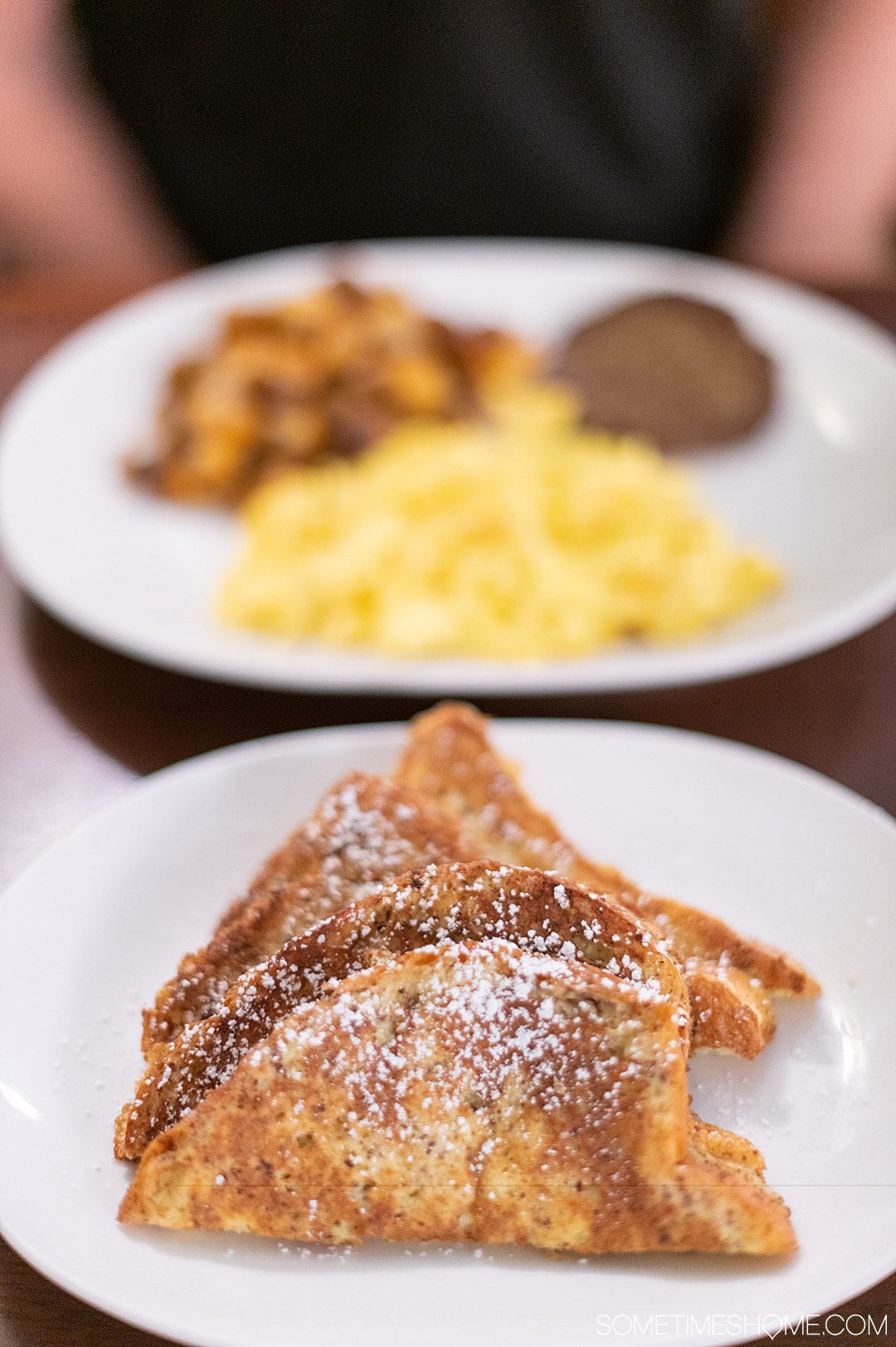 A plate of French toast in the foreground and eggs, potatoes and sausage blurry in the background.