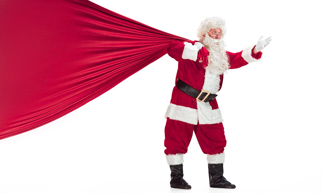 Portrait of Man in Santa Claus Costume - with a Luxurious White Beard, Santa's Hat and a Red Costume - in Full Length isolated on a White Background with big bag of gifts