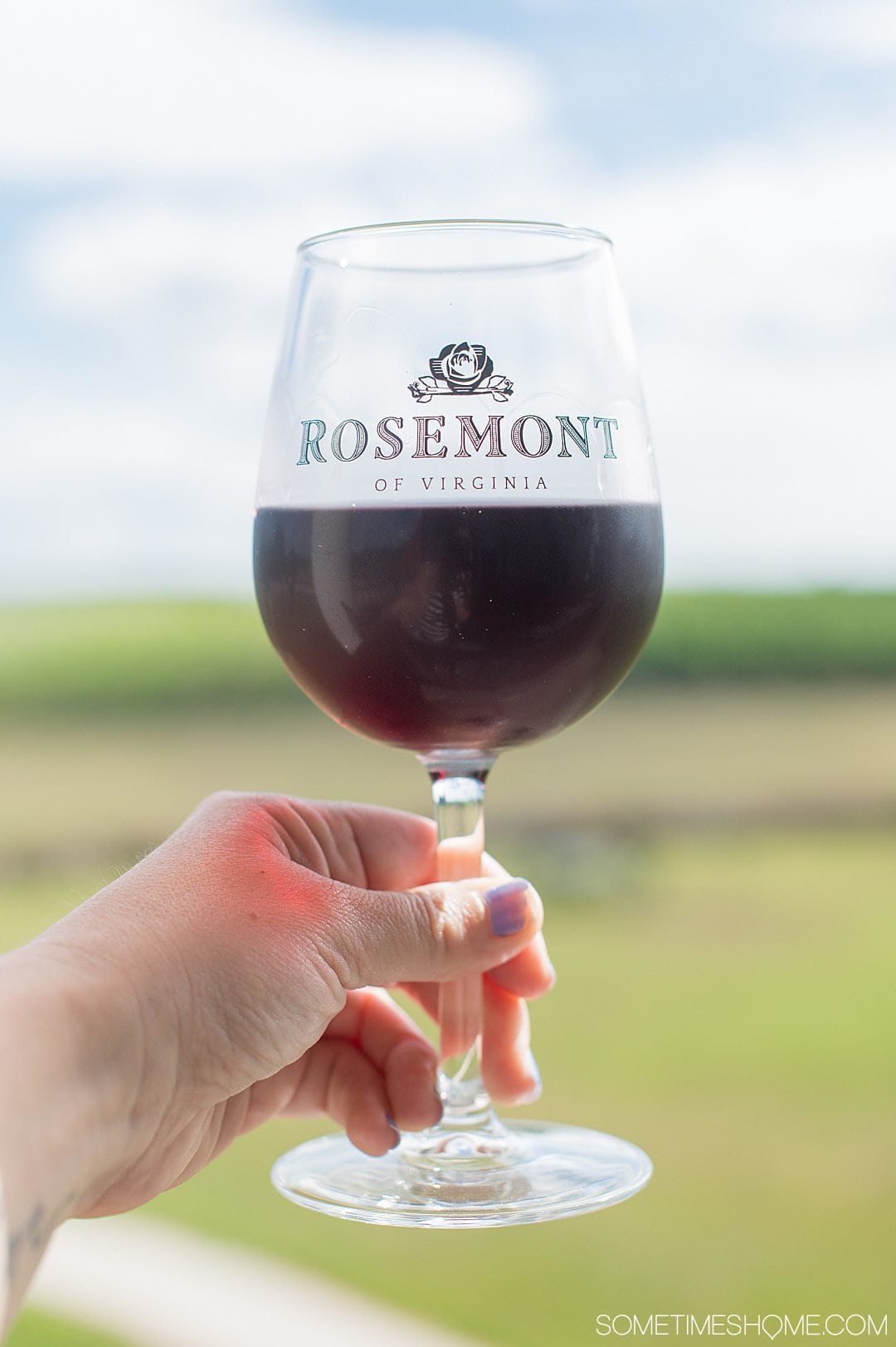 Rosemont vineyard wine glass with red wine and a woman's hand holding it against a blurry background of greenery in Virginia.