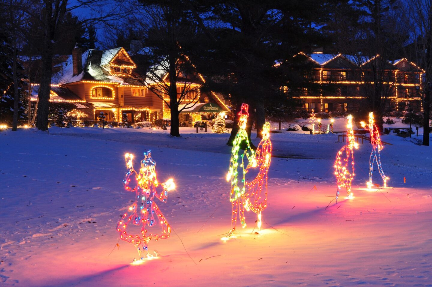 Holiday lights in the shape of people in the snow with a house with white Christmas lights in the background. Visiting Chetola Resort in Blowing Rock is one of the great winter events in North Carolina.