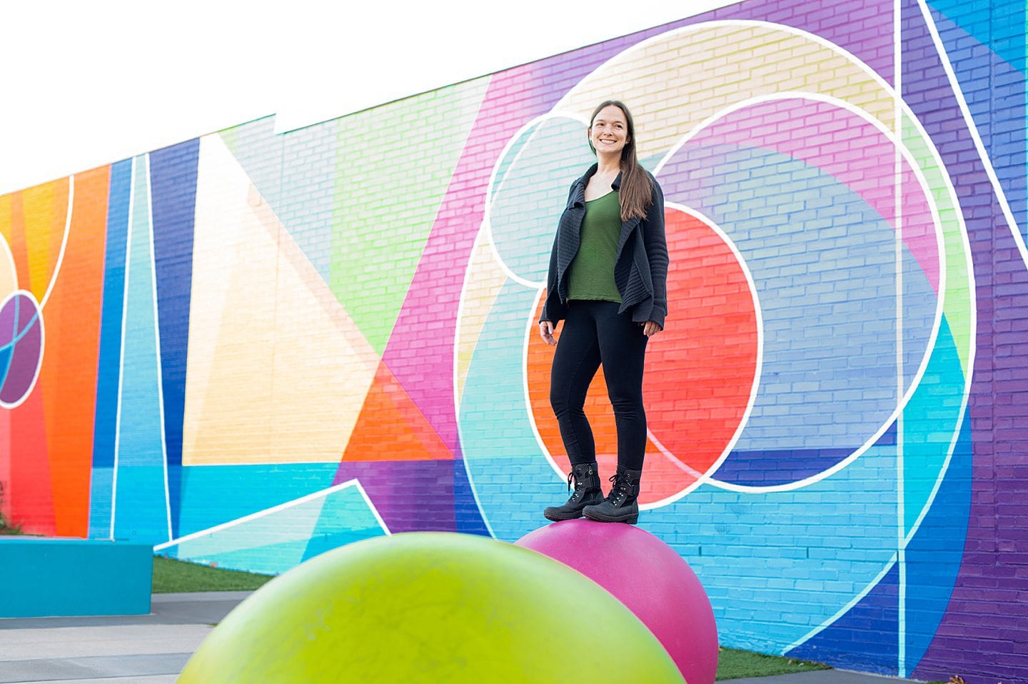 Woman on top of a large pink ball in black leggings, green top and black cardigan, with a colorful background with geometric shapes.