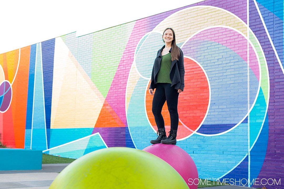 Woman on top of a large pink ball in black leggings, green top and black cardigan, with a colorful background with geometric shapes.