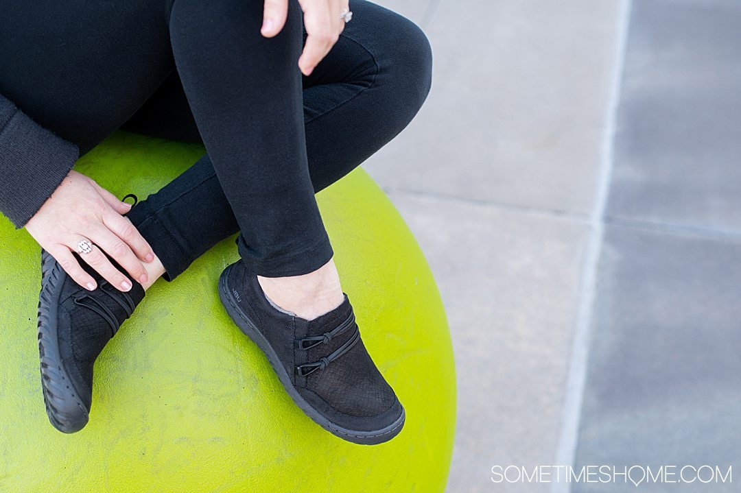 Woman's crossed legs with black Jambu & Co. Ashton shoes on a green background.