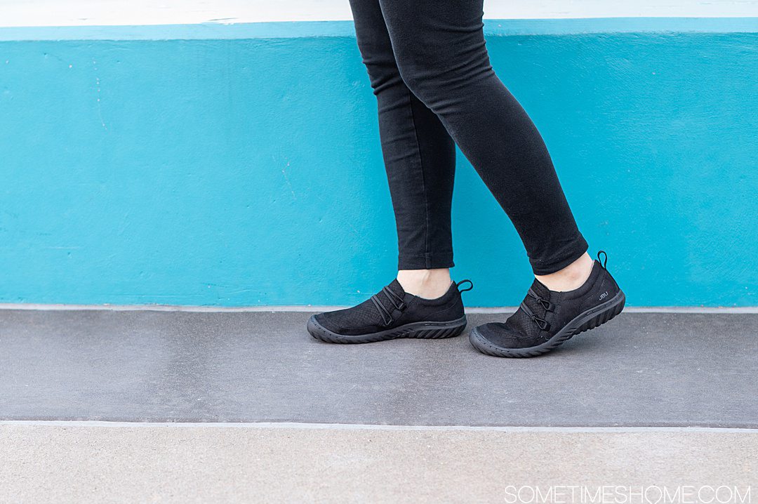 Black pair of women's flat Jambu shoes against a blue background and sidewalk for a review.