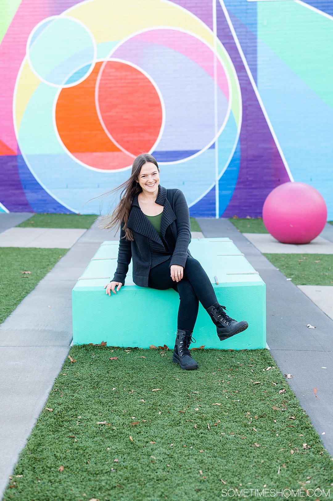 Woman sitting on a mint green bench in black leggings jeans and a black cardigan with black combat-boot inspired shoes on from Jambu & Co. and a colorful mural in the background.