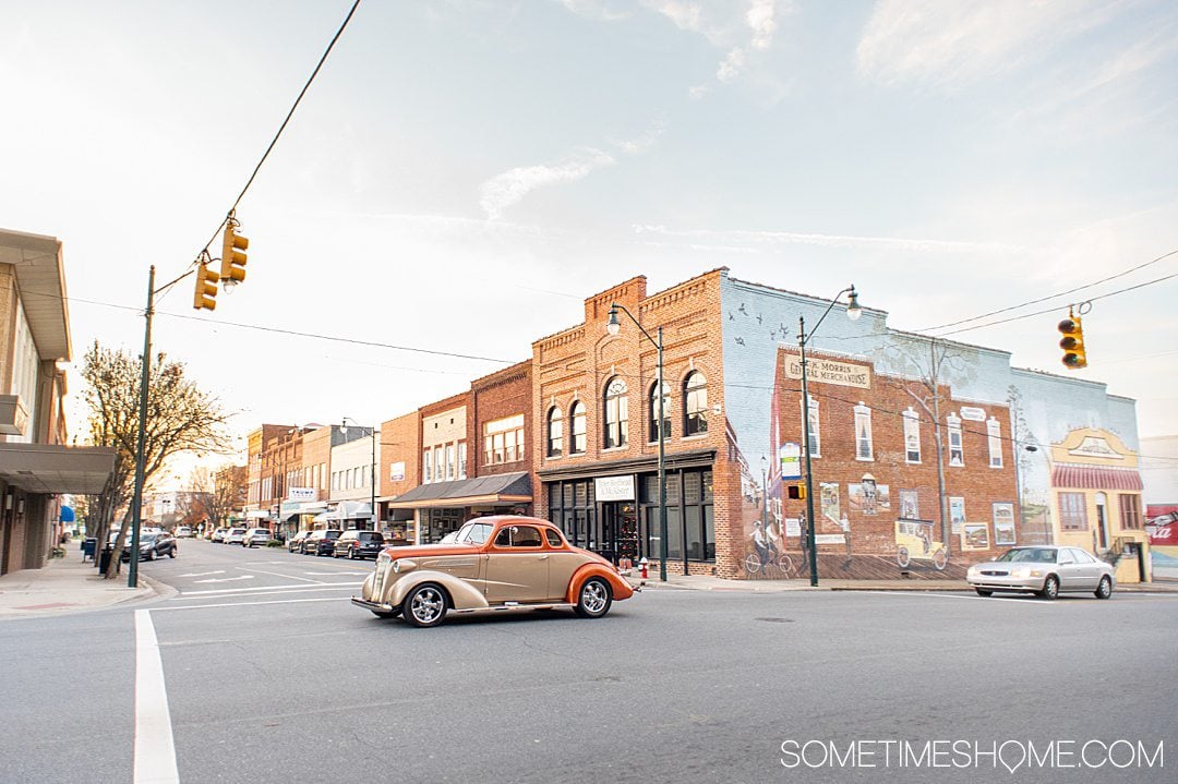 A vintage car drives past a downtown street with a mural on the side of a building in Asheboro, NC.