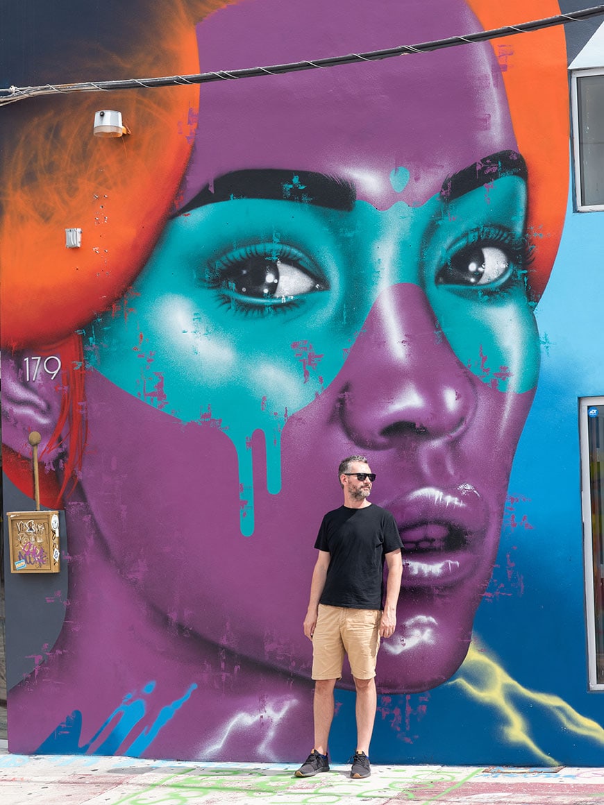 Man in front of a mural in purple, teal and orange of a woman's face in Wynwood.