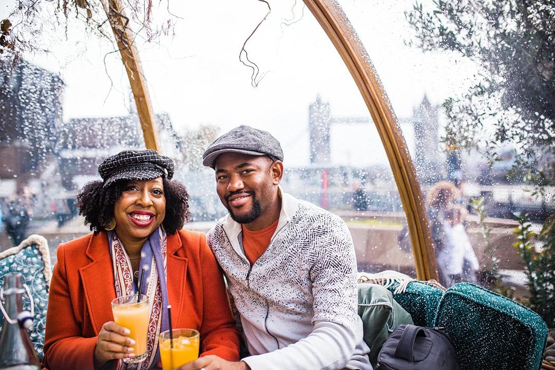 Couple inside a clear bubble with rain on it. They're holding two glasses of orange juice.