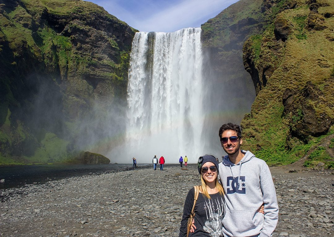 Couple in the foreground to the right of the image with a waterfall and rainbow in the background.