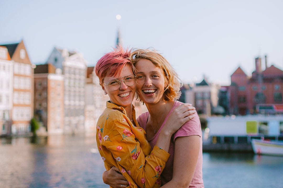 Couple travel bloggers, females side-by-side, one in yellow with pink hair and the other in a pink t-shirt with blond hair.