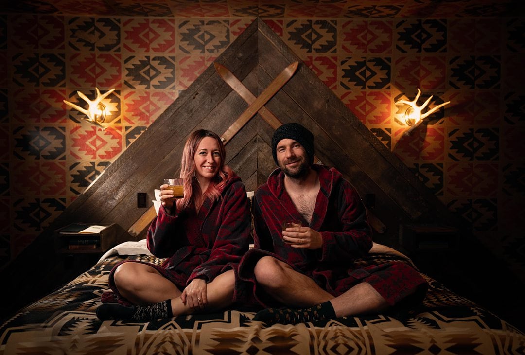 Couple sitting cross-legged on a bed in dark red bathrobes in a woodsy-feeling, darkly lit room.