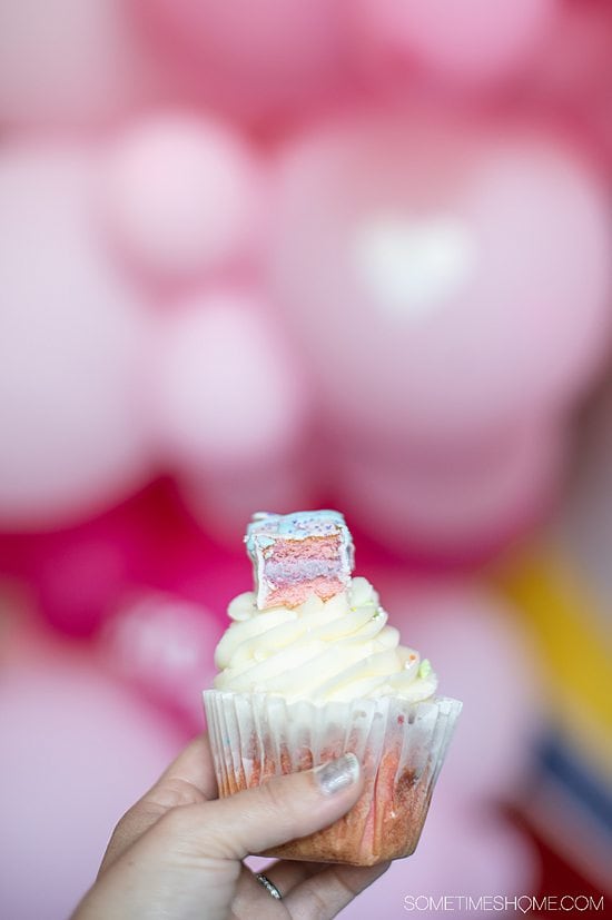 A white frosted cupcake on pink cake wth a pink and purple cookie on top, in front of blurry pink balloons.