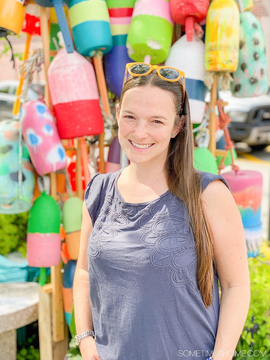 Woman in a grey shirt with yellow sunglasses on top of her head, with colorful lobster buoys behind her.