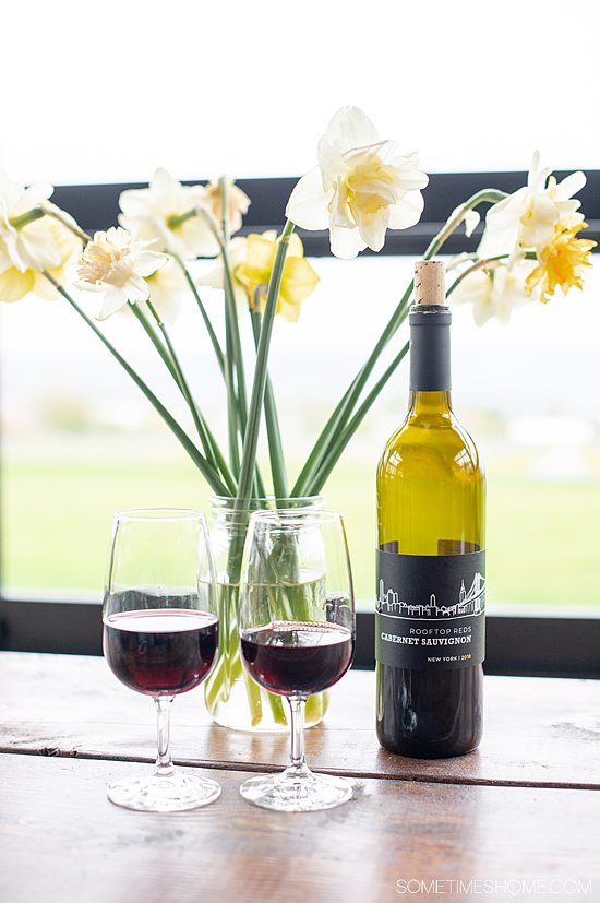 Two glasses of red wine with a wine bottle next to it and a vase of yellow and green daffodils.