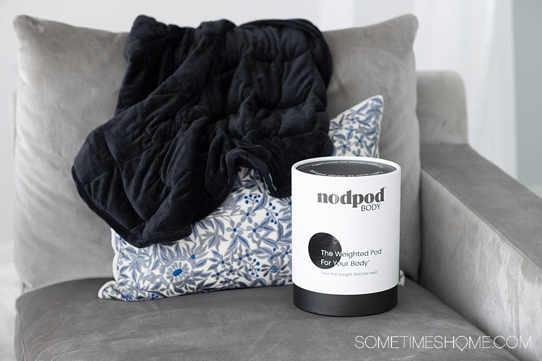 nodpod washable weighted blanket packaging in blank and white, on a grey couch with a black weighted blanket & throw pillow.