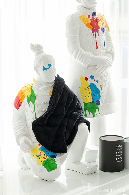White statue with rainbow splatter, and a black weighted blanket over a shoulder with a black cylindrical container nearby.