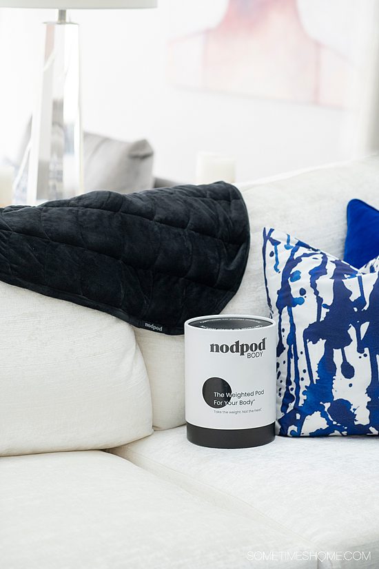 Black blanket on the back of a white couch with blue and white throw pillows and a cylindrical nodpod container.