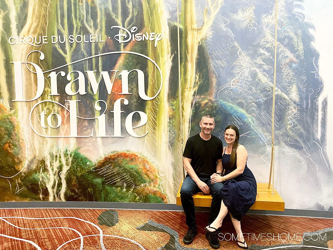 Couple on a swing in front of a 2D backdrop of a forest, with "Drawn to Life" words to the left at Cirque du Soleil.