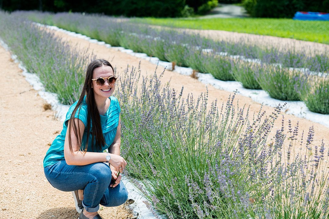 Woman with sunglasses on, smiling crouched down next to rows of lavender flowers.