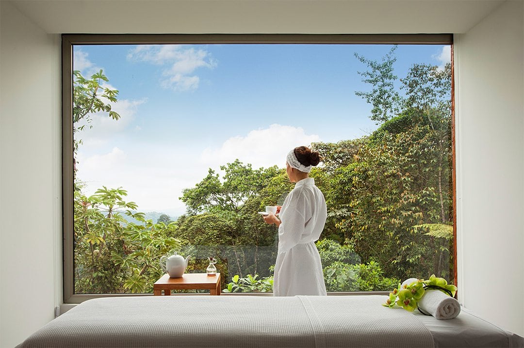 Woman looking out the window in a bathrobe holding tae, at the tropical forest at Mashpi Lodge. 