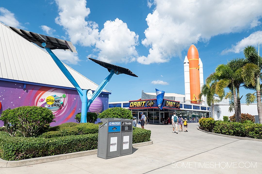 Rocket in the distance and purple kids play building in the foreground at Kennedy Space Center, with blue skies and white clouds.