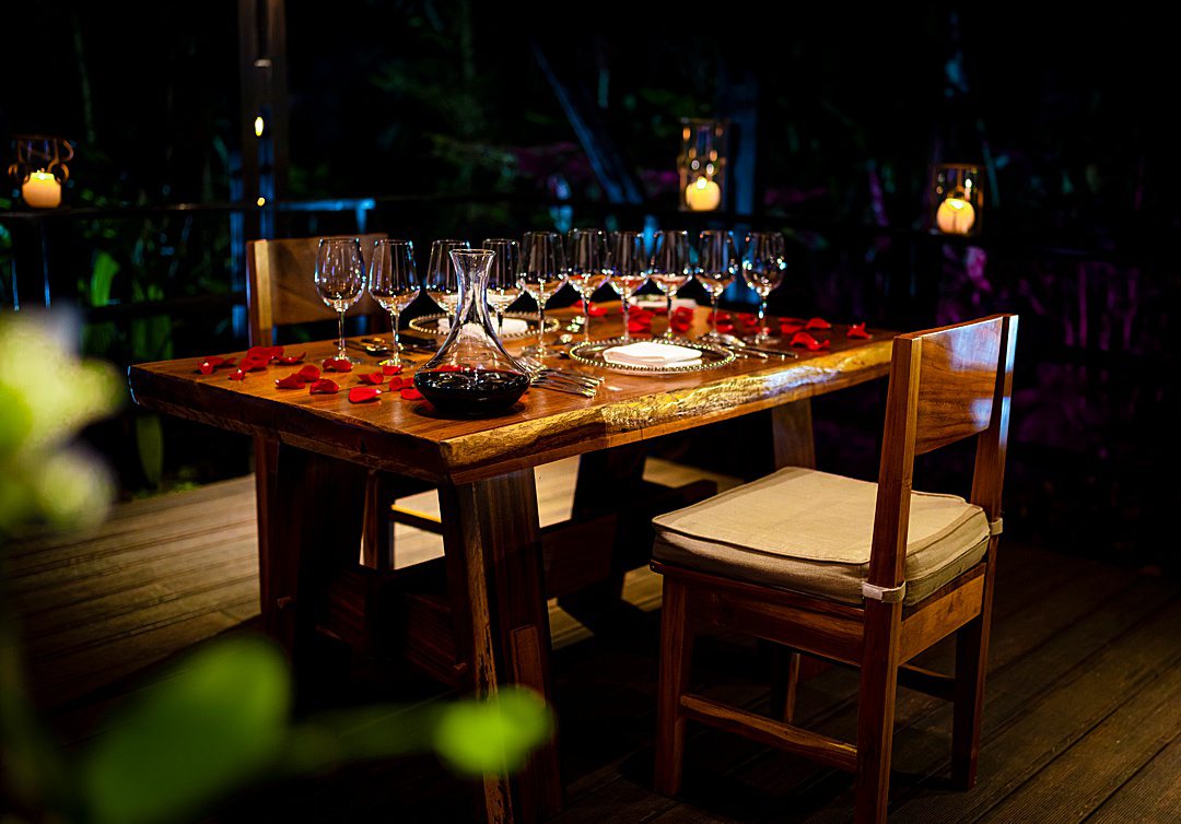 Rectangular dining table outside in a jungle oasis in the dark with a row of wine glasses in the center, rose petals and two place settings at Tabacon Resort & Spa for a private dinner.
