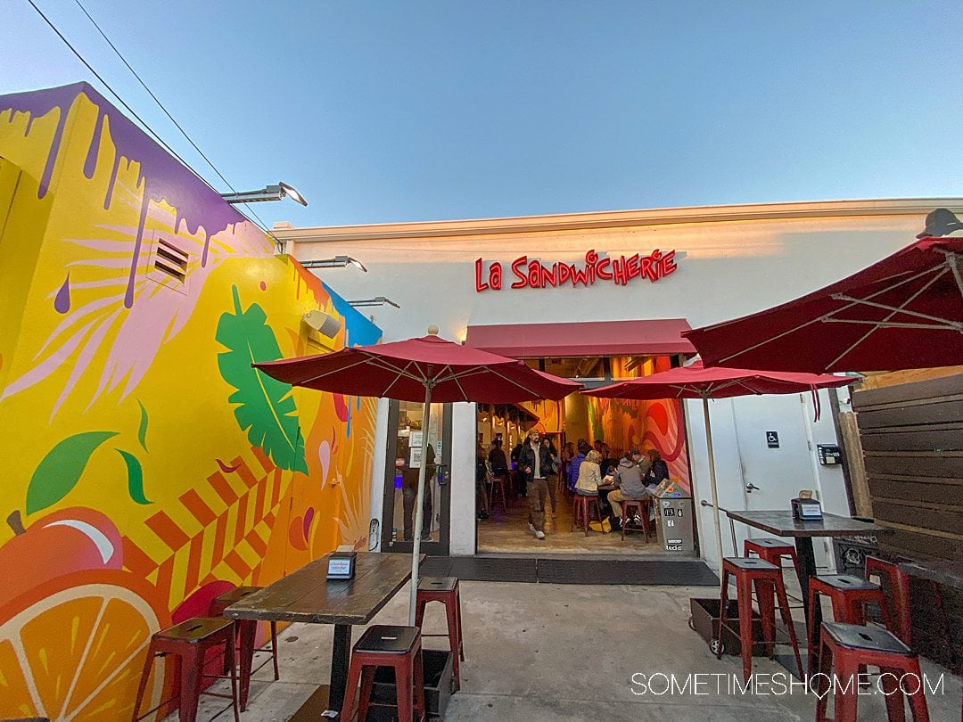 Colorful exterior of a restaurant with red umbrellas and a red sign reading, "Le Sandwicherie." 