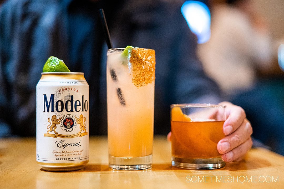 Three drinks in a row: Modelo beer can with a lime wedge on top, a Paloma in a tall glass, and hand on a rocks glass with a Mole Old Fashioned at Lola Gaspar in Santa Ana.