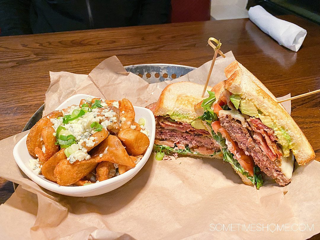 Small white bowl of fries topped with cheese and green onions and a hamburger sandwich cut in half on the right from Cin Cin in Winston-Salem.