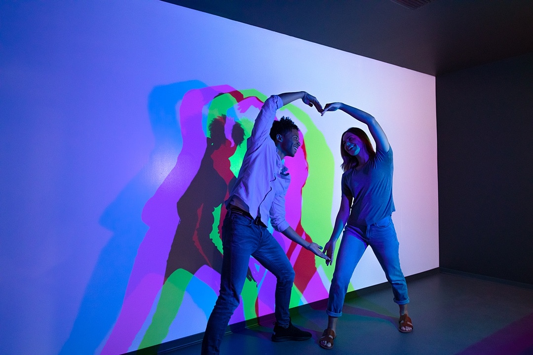 Two people making a heart with their arms against a white wall, with colorful shadows at the Museum of Illusions in Orlando, Florida.